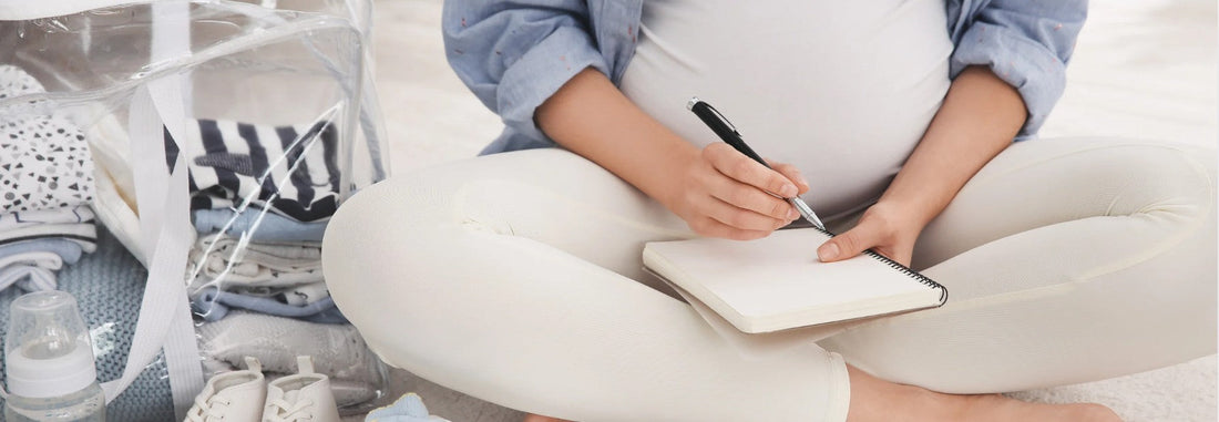 A woman on the floor, jotting notes in a notebook, preparing a list for her maternity bag essentials.