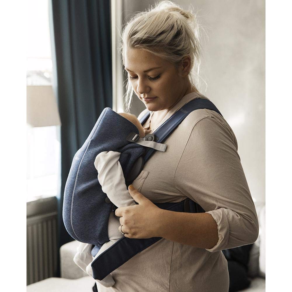 Baby Bjorn Baby Carrier Mini in navy blue carried by woman with young infant in facing in position close to her heart while holding infant's hand